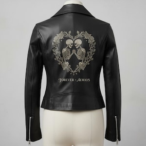 Puede incluir: Chaqueta de cuero negra con cremalleras plateadas. La espalda presenta un diseño en forma de corazón con dos esqueletos tomados de la mano, rodeados de rosas y la frase "Forever + Always".