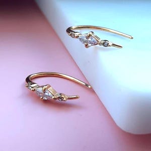 May include: Two gold-coloured hoop earrings. Each earring features a marquise-cut clear gemstone and several smaller clear gemstones. The earrings are displayed on a pink and white surface.