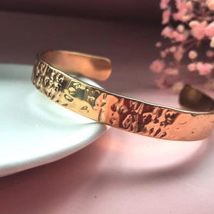 May include: A polished rose gold-toned metal cuff bracelet featuring a hammered, textured surface. The bracelet has an open-ended design with rounded edges, reflecting light across its uneven, dimpled exterior finish.