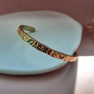 May include: A gold-toned metal cuff bracelet featuring a textured, hammered surface with irregular indentations. The bracelet has an open-ended design, showcasing a reflective, metallic finish that catches the light across its uneven, organic-looking exterior.