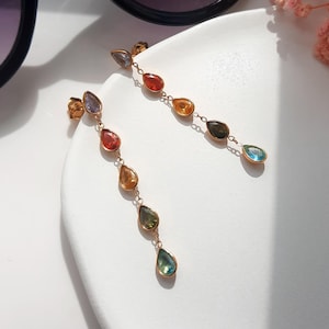 May include: A pair of long drop earrings featuring a gold-tone chain with multiple teardrop-shaped gemstones in various colours, including red, orange, yellow, green, and blue, suspended in a vertical arrangement from a small gold stud post.