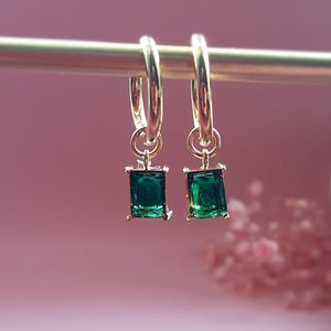 May include: A pair of gold hoop earrings, each featuring a dangling emerald green rectangular gemstone charm. The earrings are suspended from a thin gold bar, showcasing a polished metallic finish and vibrant green faceted stones in a simple setting.