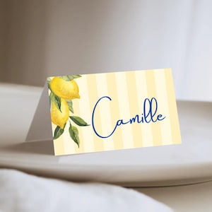 Peut inclure: Une carte de place pliée avec un fond à rayures jaunes et blanches. La carte présente une illustration à l'aquarelle de citrons et de feuilles. Le nom "Camille" est écrit en bleu.