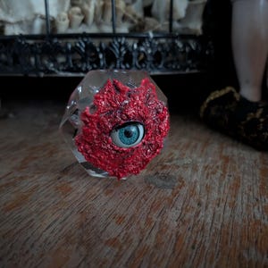 Puede incluir: Un objeto de vidrio transparente y facetado con una forma orgánica roja y texturizada en un lado, con un ojo azul. El objeto descansa sobre una superficie de madera. El tema general es gótico o macabro.