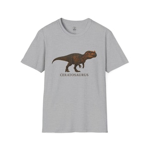 May include: A light gray t-shirt featuring a detailed illustration of a Ceratosaurus dinosaur in shades of brown and orange. The dinosaur image is centered on the shirt, with the name "CERATOSAURUS" printed below it in a simple font.