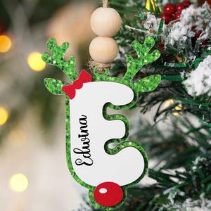 May include: A Christmas ornament shaped like the letter "E" with a reindeer design. The green glitter antlers are paired with a red bow and nose. The name "Edwina" is written in black script. The ornament hangs from a wooden bead string.