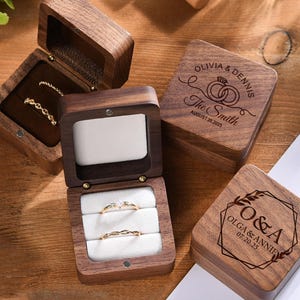 Può includere: Scatole per anelli in legno, alcune aperte per mostrare anelli d'oro con disegni intricati e un diamante. Le scatole sono incise con nomi e date, perfette per matrimoni o anniversari. Le scatole sono in legno scuro.