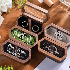 May include: Hexagonal wooden ring boxes with clear lids, displaying rings and engraved names and dates. The boxes are arranged on a wooden surface. Examples of engraved text include "Emma & Matt 02.12.2025" and "David+Linda 09.20.2024".