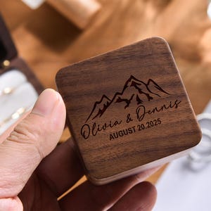 May include: A square, wooden ring box featuring a mountain range design and the names "Olivia & Dennis" with the date "AUGUST 20.2025" engraved on the lid. The box is held in a hand, with a partially visible open ring box in the background.