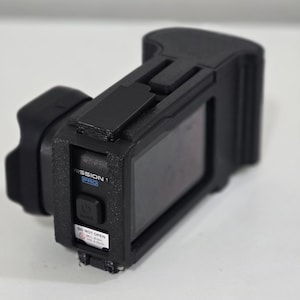 Peut inclure: Un bo&icirc;tier de protection noir pour cam&eacute;ra d'action avec un cadre textur&eacute;, un bouton d'alimentation et une &eacute;tiquette portant l'inscription MISSION 1 PRO. Un petit autocollant blanc d'avertissement indique DO NOT OPEN Mic drain, not a door.