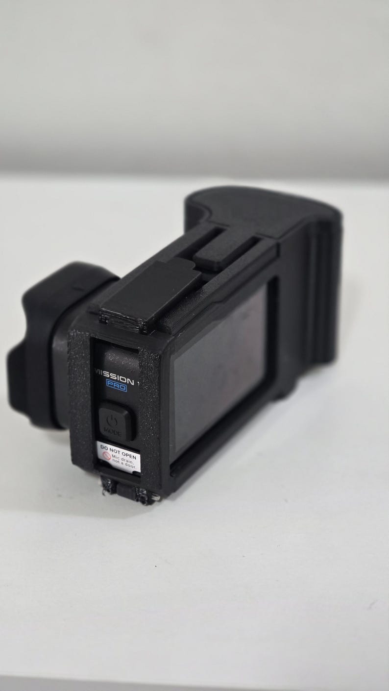 Peut inclure: Un bo&icirc;tier de protection noir pour cam&eacute;ra d'action avec un cadre textur&eacute;, un bouton d'alimentation et une &eacute;tiquette portant l'inscription MISSION 1 PRO. Un petit autocollant blanc d'avertissement indique DO NOT OPEN Mic drain, not a door.