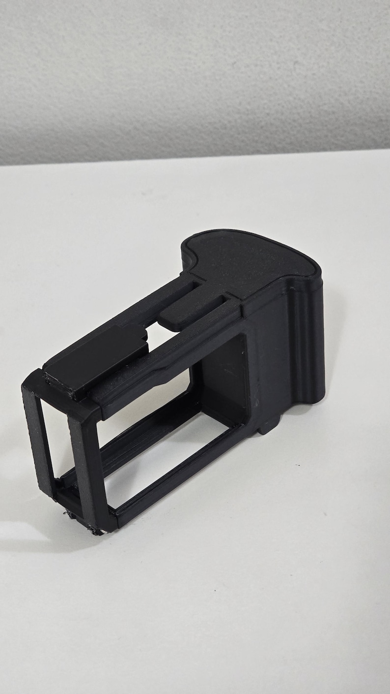 Peut inclure: Un cadre ou bo&icirc;tier de protection noir, rectangulaire et imprim&eacute; en 3D pour un petit appareil &eacute;lectronique. L'objet pr&eacute;sente une conception en cage &agrave; c&ocirc;t&eacute;s ouverts avec une section de poign&eacute;e solide et profil&eacute;e &agrave; une extr&eacute;mit&eacute;.
