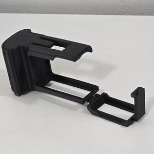 Peut inclure: Un cadre de protection pour cam&eacute;ra imprim&eacute; en 3D de couleur noire, dot&eacute; d'un bo&icirc;tier rectangulaire avec un dessus ouvert et une porte lat&eacute;rale articul&eacute;e. La structure comprend une poign&eacute;e solide &agrave; une extr&eacute;mit&eacute; et un cadre rigide.
