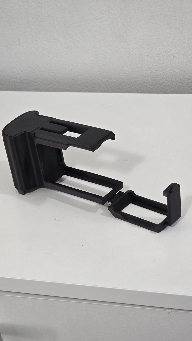 Peut inclure: Un cadre de protection pour cam&eacute;ra imprim&eacute; en 3D de couleur noire, dot&eacute; d'un bo&icirc;tier rectangulaire avec un dessus ouvert et une porte lat&eacute;rale articul&eacute;e. La structure comprend une poign&eacute;e solide &agrave; une extr&eacute;mit&eacute; et un cadre rigide.