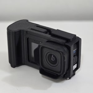 Peut inclure: Une cam&eacute;ra d'action noire log&eacute;e dans un bo&icirc;tier de protection avec une poign&eacute;e int&eacute;gr&eacute;e sur le c&ocirc;t&eacute; gauche. La cam&eacute;ra pr&eacute;sente un objectif carr&eacute;, un petit &eacute;cran d'affichage frontal et un bouton d'alimentation lat&eacute;ral avec le texte SION visible.