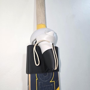 Op de afbeelding: Een cricketbat met een zwart-geel ontwerp, een witte grip en een wit en zwart object bevestigd. Op de bat staat het woord "MASURI". De grip is beige en geel.