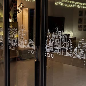 Peut inclure: Une porte vitrée ornée d'un autocollant représentant un village d'hiver blanc. L'autocollant présente des maisons, des arbres et un bonhomme de neige. La porte est encadrée de noir et reflète l'intérieur d'une pièce avec des lumières.
