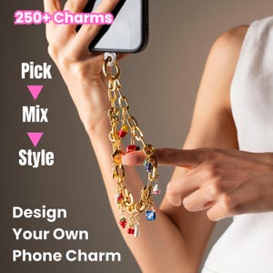May include: A gold-toned metal chain phone charm featuring various colorful miniature enamel charms including a strawberry, cherries, and a cupcake. The text 250+ Charms, Pick, Mix, Style, and Design Your Own Phone Charm is visible on the image.