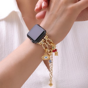 Op de afbeelding: Een goudkleurige metalen horlogeband voor een smartwatch met een schakelontwerp. De band is versierd met diverse bungelende bedels, waaronder een paar rode kersen, een blauw-wit boze oog, een kleine blauwe steen en een letter M-bedel.