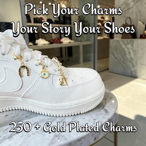 Peut inclure: Une basket blanche avec trois breloques dorées attachées aux lacets : un fer à cheval, un œil protecteur et un cœur. Le texte Pick Your Charms Your Story Your Shoes et 250 + Gold Plated Charms est visible en surimpression sur l'image.