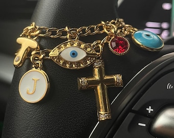 May include: A gold-tone chain charm bracelet featuring multiple pendants including a letter T, a letter J on a white circular disc, an eye-shaped charm with a blue center, a red faceted crystal, a gold cross, and a round blue evil eye charm.