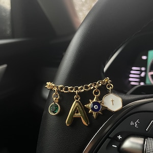 Op de afbeelding: Een goudkleurige kettingarmband met vier verschillende bedels: een ronde groene edelsteen, een gouden letter A, een stervormige blauw-witte boze oog bedel en een witte zeshoekige bedel met een klein gouden kruissymbool in het midden.