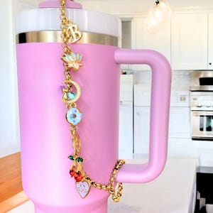 Könnte beinhalten: Ein rosa isolierter Becher mit einem goldenen Charm-Armband. Das Armband hat verschiedene Charms, darunter einen Buchstaben "B", eine Lotusblume, einen Mond, einen Donut, Kirschen und ein Herz mit dem Text "I'm good".