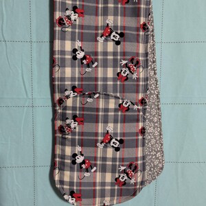 May include: A double oven mitt featuring a grey, cream, and red plaid pattern with a repeating print of Minnie Mouse. The mitt has a hanging loop at the top and a contrasting grey and white floral fabric lining visible on the side edge.
