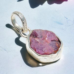 Könnte beinhalten: Silberanhänger mit einem rohen, rosafarbenen Edelstein. Der Stein ist in einer detaillierten Silberfassung mit einer strukturierten Öse gefasst. Der Anhänger wird auf einer hellblauen Oberfläche mit einer rosa Blume im Hintergrund präsentiert.