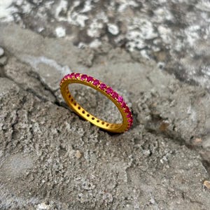 Könnte beinhalten: Ein goldfarbener Ring mit einer Reihe kleiner, leuchtend rosa Edelsteine. Der Ring ist auf einer strukturierten, grauen Oberfläche platziert, was den Kontrast zwischen dem Glanz des Rings und dem rauen Hintergrund hervorhebt. Ein Schmuckstück.