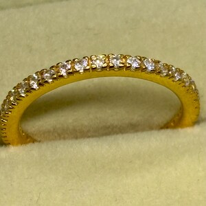 Könnte beinhalten: Ein goldfarbener Ring mit einem Band, das vollständig mit kleinen, klaren Edelsteinen besetzt ist. Der Ring ist vor einem weichen, hellgelben Hintergrund platziert, der sein Funkeln und Design hervorhebt. Die Edelsteine sind gleichmäßig angeordnet.