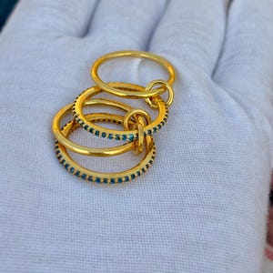 Könnte beinhalten: Eine Sammlung goldfarbener Ringe. Ein Ring ist schlicht, während die anderen mit kleinen, blauen Edelsteinen verziert sind. Die Ringe sind ineinander verschlungen und bilden ein einzigartiges Design. Die Ringe werden auf einer weißen Oberfläche präsentiert.