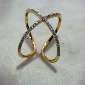 Könnte beinhalten: Ein goldfarbener Ring mit einem Kreuzmuster. Der Ring hat ein Band aus kleinen, klaren Steinen entlang der Oberseite des Kreuzes und eine glatte, polierte Oberfläche auf dem Rest des Bandes. Der Ring hat ein modernes, offenes Design.