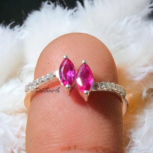 Könnte beinhalten: Ein silberner Ring mit zwei leuchtend rosa, marquiseförmig geschliffenen Edelsteinen. Das Band ist mit kleinen, klaren Steinen besetzt. Das Wort "Siddharth" ist auf dem Ringband eingraviert. Der Ring wird vor einem weichen, weißen Hintergrund gezeigt.