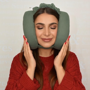 Könnte beinhalten: Ein salbeigrünes Reisekissen zur Unterstützung von Kopf und Nacken. Das Kissen hat eine gebogene Form und einen Riemen zur Befestigung. Die Person im Bild trägt einen roten Pullover und hält das Kissen.