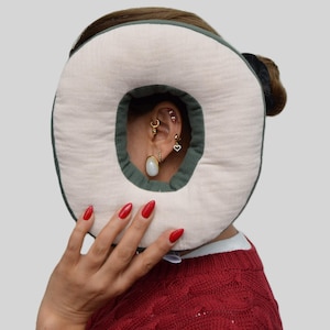 Puede incluir: Una almohada para el cuello de color crema con un borde verde se sostiene frente a la cara de una persona. La persona lleva un suéter rojo y tiene múltiples aretes. Se ve la mano de la persona con esmalte de uñas rojo.