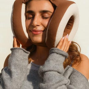 Puede incluir: Una máscara para dormir marrón y crema es usada por una persona con los ojos cerrados. La máscara está hecha de material suave y cubre los ojos y las orejas. La persona lleva un suéter gris.