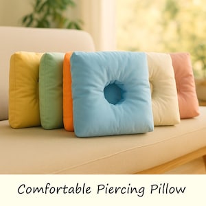 May include: A selection of square pillows in shades of yellow, green, orange, blue, and peach. The blue pillow features a central hole. The pillows are placed on a beige sofa. The text "Comfortable Piercing Pillow" is displayed below.