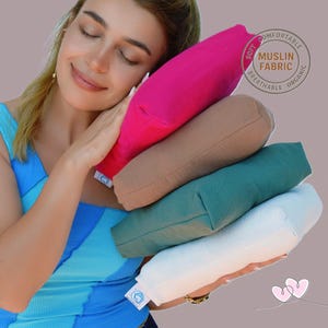 May include: A stack of four colorful, oblong-shaped pillows in shades of pink, tan, teal, and white. The pillows are made of soft muslin fabric, as indicated by a circular label. The pillows are being held by a person wearing a blue top.