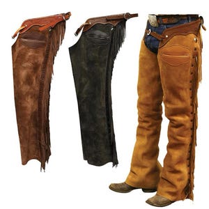 Op de afbeelding: Drie paar leren chaps met franjes. De chaps zijn bruin, zwart en beige. De bruine en zwarte chaps hangen, terwijl de beige chaps door een persoon worden gedragen.