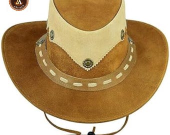 May include: A brown suede cowboy hat with a cream-colored crown and decorative stitching. The hat features a chin strap and metal accents. The brim is wide and the hat has a classic Western style.