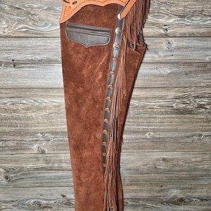 May include: Brown leather chaps with fringe detail and a brown suede panel. The chaps have a belt loop and a buckle closure.
