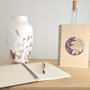 Puede incluir: Un jarr&oacute;n de cer&aacute;mica blanca con un dise&ntilde;o floral en tonos marr&oacute;n y melocot&oacute;n. Un cuaderno de espiral con una cubierta de madera y un dise&ntilde;o de flor recortada, una botella de vidrio transparente y un cuaderno abierto con un bol&iacute;grafo sobre una superficie de madera.