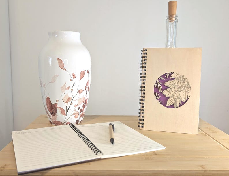 Puede incluir: Un jarr&oacute;n de cer&aacute;mica blanca con un dise&ntilde;o floral en tonos marr&oacute;n y melocot&oacute;n. Un cuaderno de espiral con una cubierta de madera y un dise&ntilde;o de flor recortada, una botella de vidrio transparente y un cuaderno abierto con un bol&iacute;grafo sobre una superficie de madera.