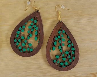 May include: Teardrop-shaped wooden earrings with a dark brown frame and a green leaf design. The earrings have gold-colored ear wires. The earrings are displayed on a light brown wooden surface.