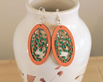May include: Oval-shaped wooden earrings with an orange frame and a green leaf design. The earrings are hanging from silver hooks. The earrings are displayed on a white vase.