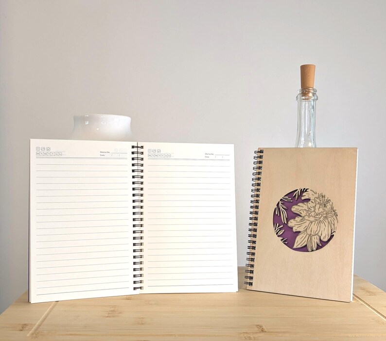 Puede incluir: Dos cuadernos con espiral sobre una superficie de madera. Uno est&aacute; abierto, mostrando p&aacute;ginas rayadas. El otro tiene una cubierta marr&oacute;n con un recorte circular que revela un fondo p&uacute;rpura y un dise&ntilde;o floral. Una botella de vidrio transparente con un tap&oacute;n de corcho est&aacute; detr&aacute;s de los cuadernos.