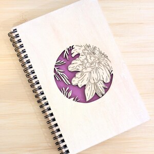 Puede incluir: Un cuaderno de espiral de madera con un dise&ntilde;o floral cortado con l&aacute;ser. La cubierta es de madera de color claro, con un recorte circular que revela un fondo p&uacute;rpura. El dise&ntilde;o floral es un contorno detallado de una flor.