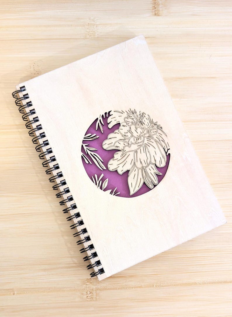 Puede incluir: Un cuaderno de espiral de madera con un dise&ntilde;o floral cortado con l&aacute;ser. La cubierta es de madera de color claro, con un recorte circular que revela un fondo p&uacute;rpura. El dise&ntilde;o floral es un contorno detallado de una flor.