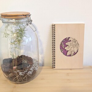 Puede incluir: Un gran terrario de vidrio con tapa de madera y cierre met&aacute;lico, que contiene tierra, rocas y vegetaci&oacute;n. Un cuaderno de espiral de madera con un dise&ntilde;o floral est&aacute; junto al terrario. El cuaderno tiene un c&iacute;rculo morado con un dise&ntilde;o floral.